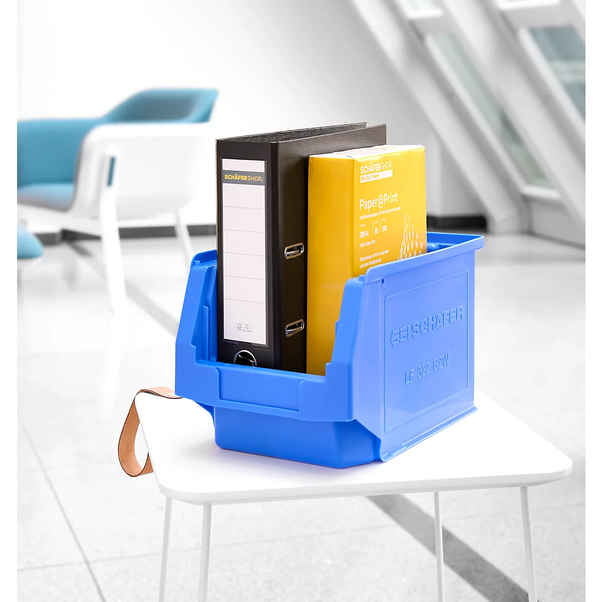 Blaue Sichtlagerbox auf einem weißen Tisch, gefüllt mit einem schwarzen Ordner und gelben Papier.