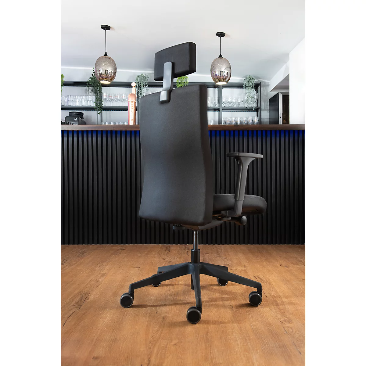 Un fauteuil de bureau noir se dresse devant un fond sombre. La chaise a un appui-tête et des roulettes.