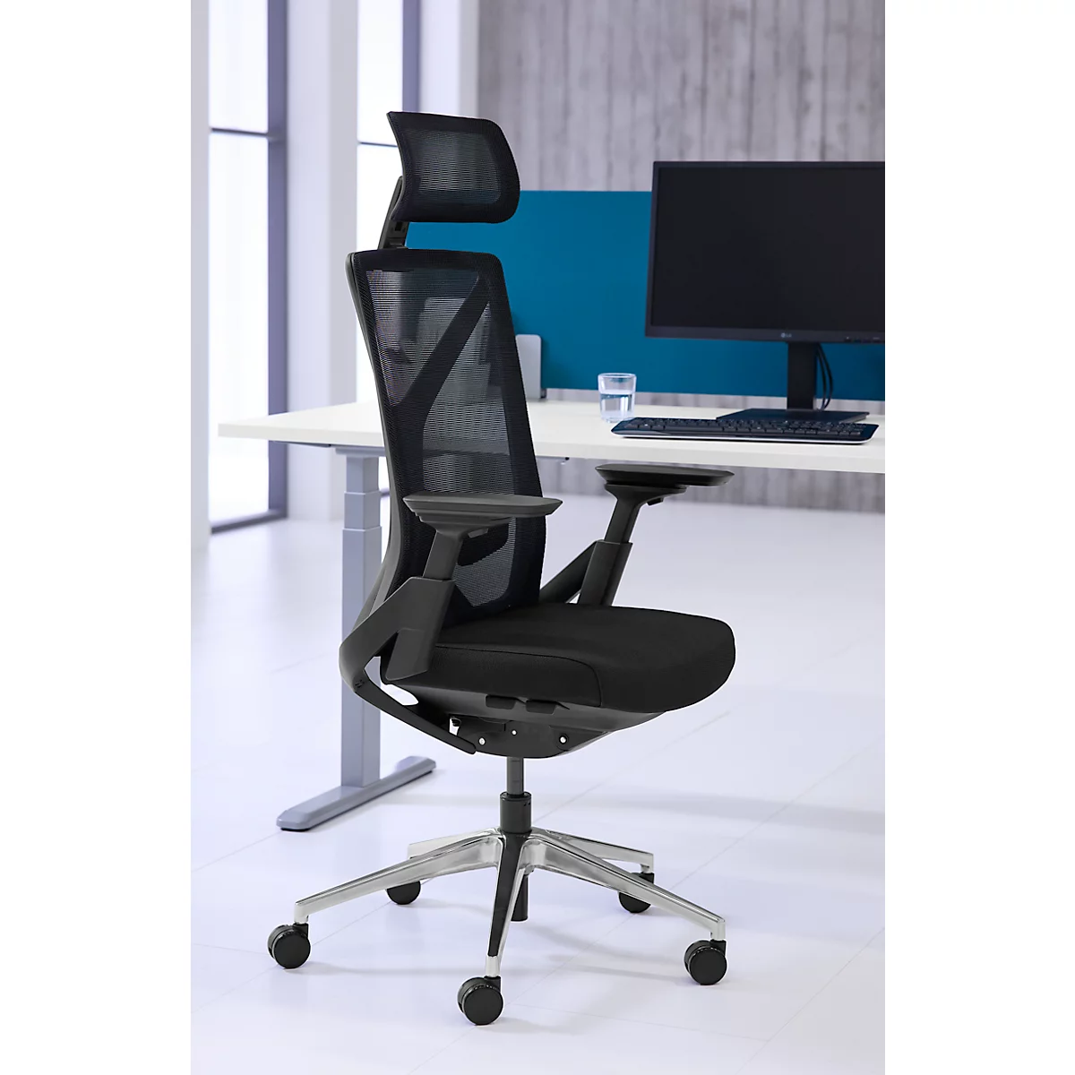 Chaise de bureau noire avec dossier en maille et appuie-tête. Elle se trouve devant un bureau avec un ordinateur. Pieds et roulettes argentés.