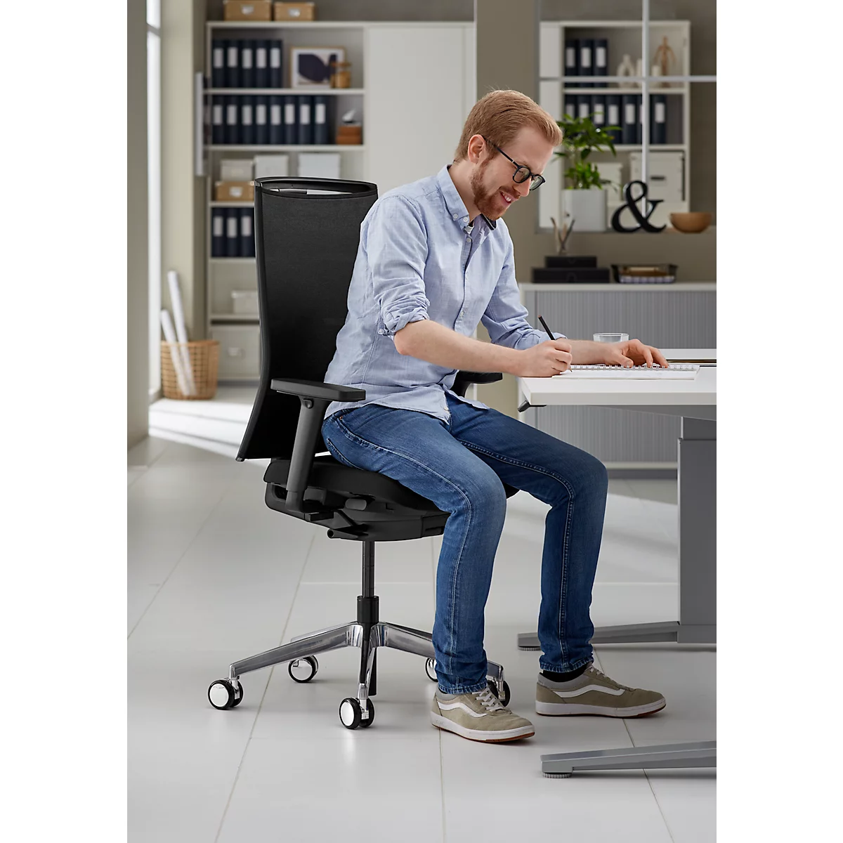 Un homme est assis dans un fauteuil de bureau noir et écrit sur une feuille de papier au bureau, en souriant.