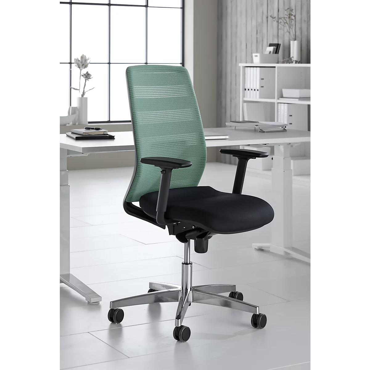 Chaise de bureau avec dossier en maille verte et assise noire, devant un bureau blanc. Fenêtre en arrière-plan.