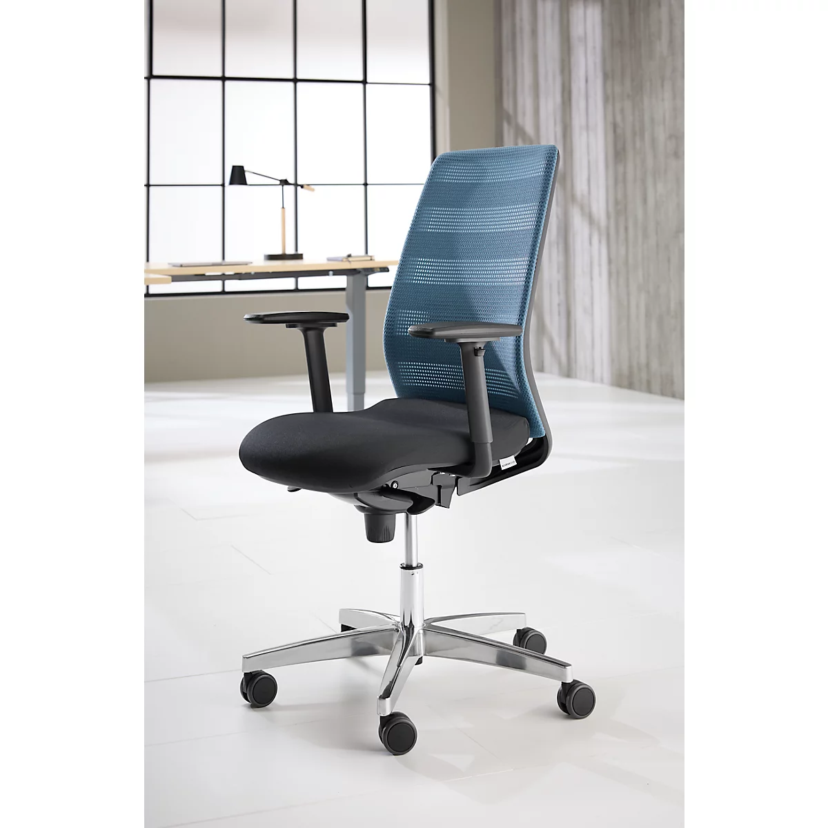 Chaise de bureau avec dossier en filet bleu et assise noire. La chaise est devant une fenêtre.