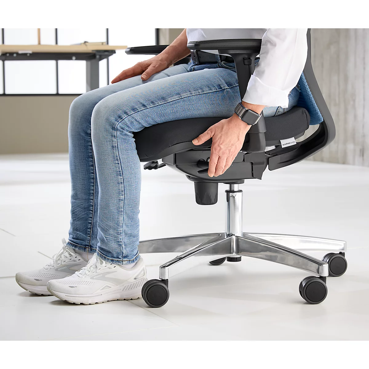 Personne assise sur une chaise de bureau noire. Personne portant un jean, une chemise blanche, une montre connectée. Bras appuyé sur la chaise. Vue sur les jambes et les pieds.
