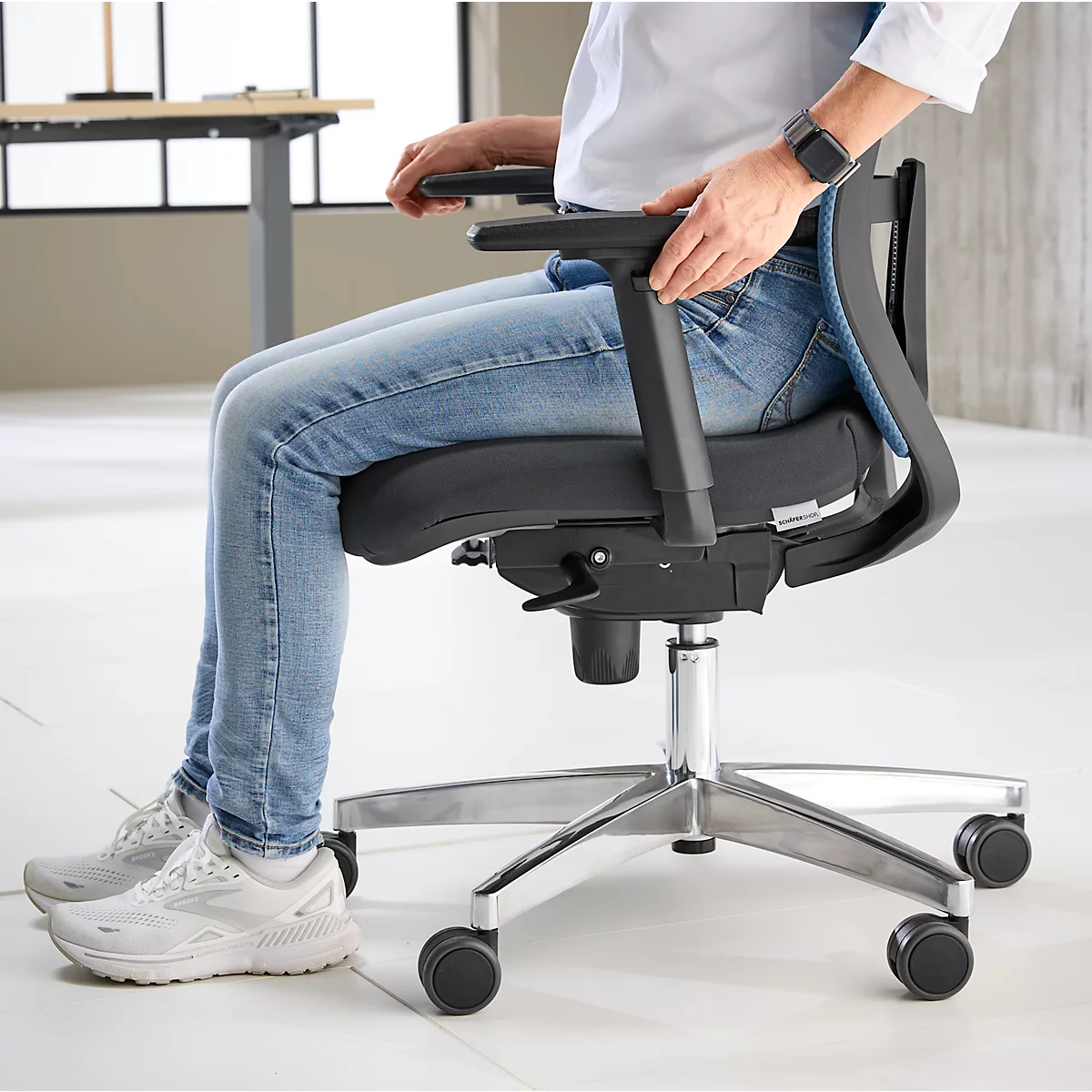 Personne assise sur une chaise de bureau. Dossier en maille bleu clair, assise noire, cadre argenté, roulettes.