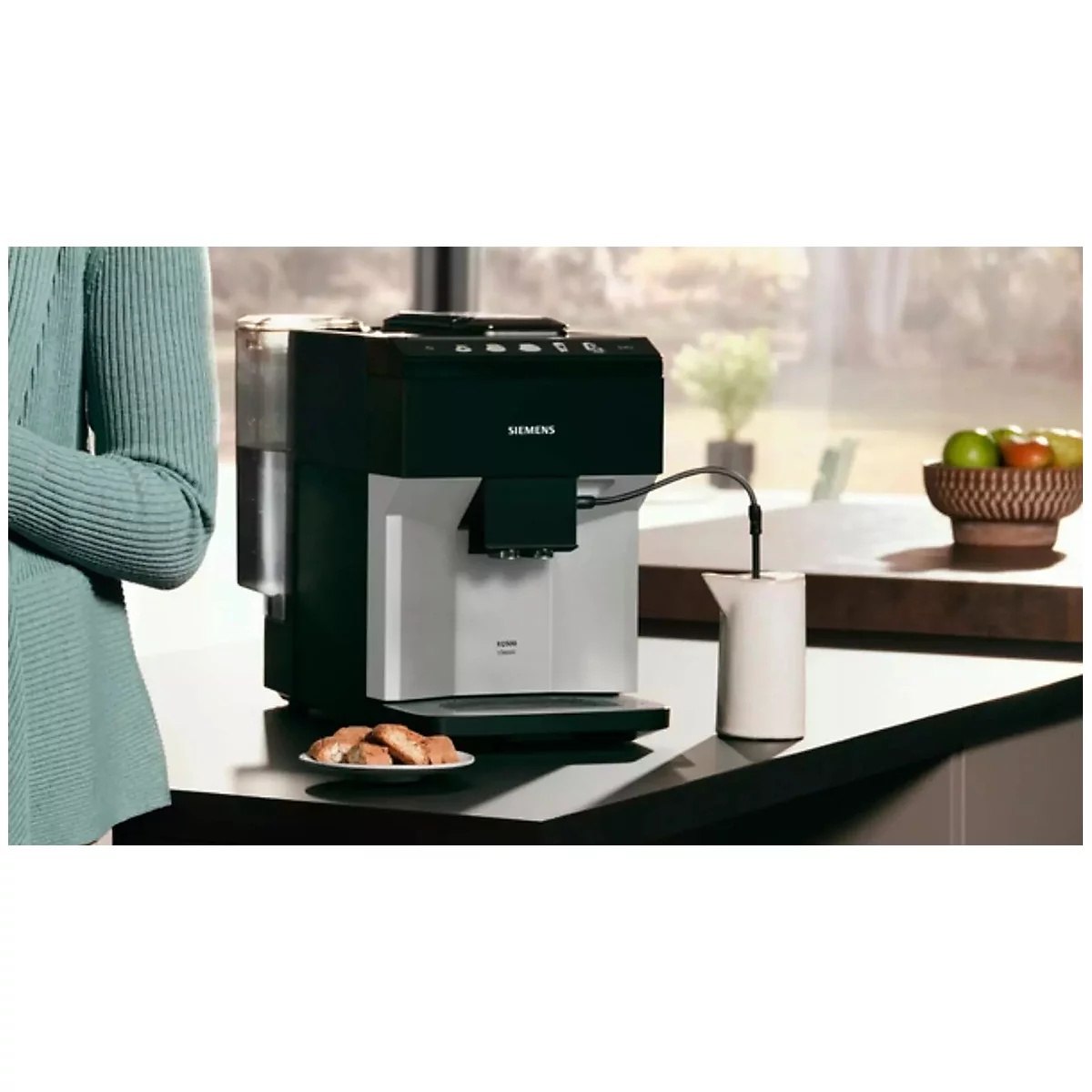 Eine Kaffeemaschine mit Milchaufschäumer. Daneben Gebäck und Obst. Im Hintergrund eine Frau.
