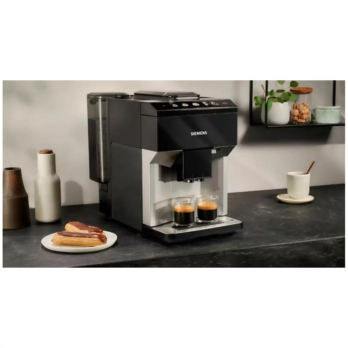Eine schwarze Kaffeemaschine mit zwei Espressogläsern. Auf dem Tisch stehen Gebäck, eine Tasse und Dekorationsartikel.