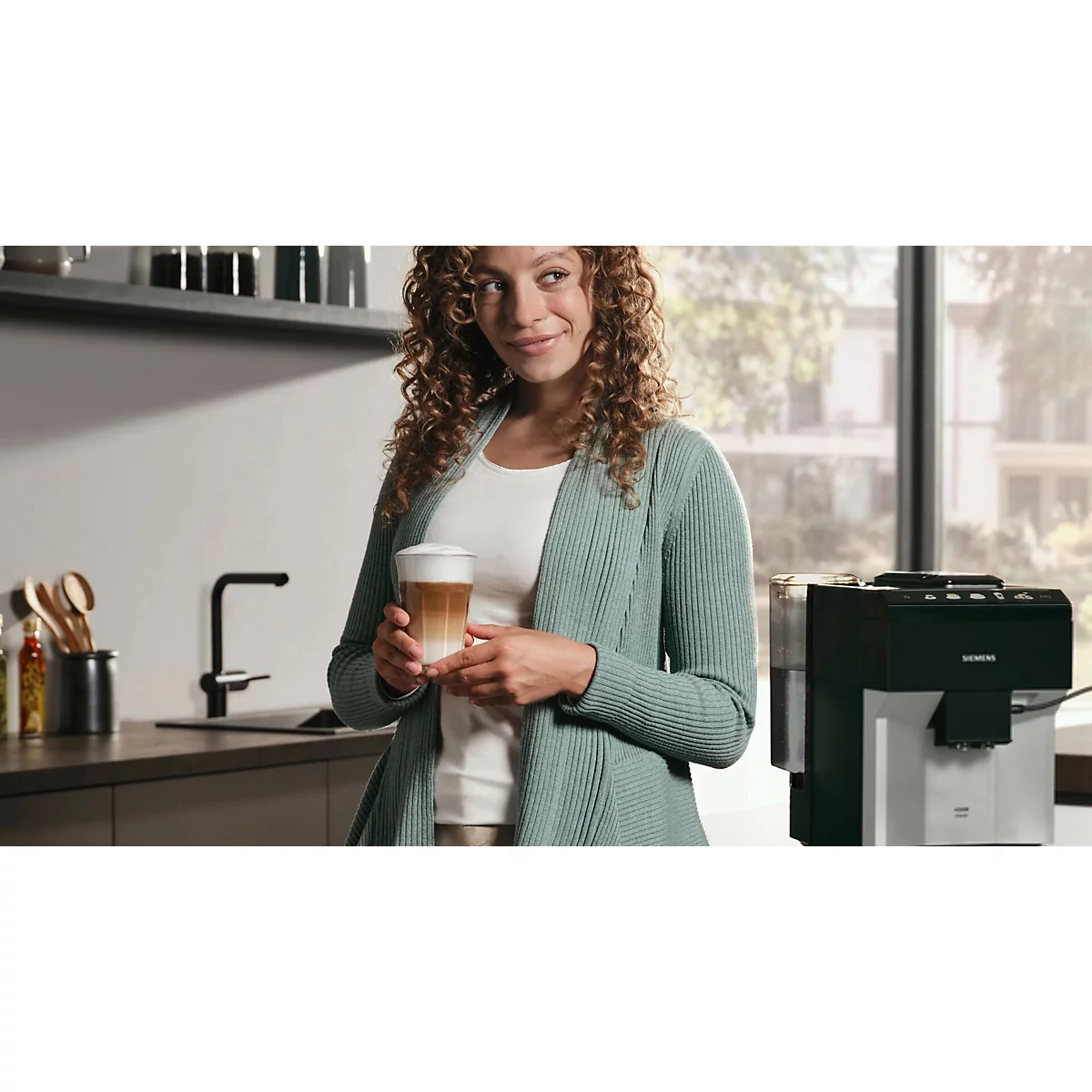Frau mit lockigem Haar hält ein Kaffeeglas, lächelt. Steht neben Kaffeemaschine in einer Küche.
