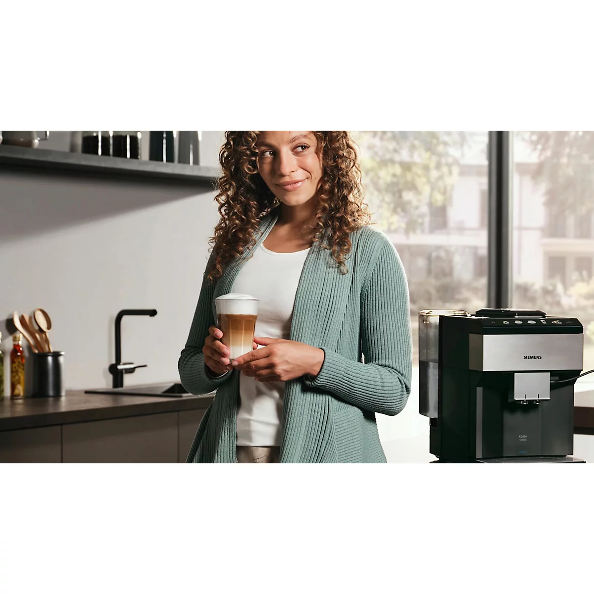 Frau hält einen Latte Macchiato in der Küche, Kaffeemaschine im Hintergrund.