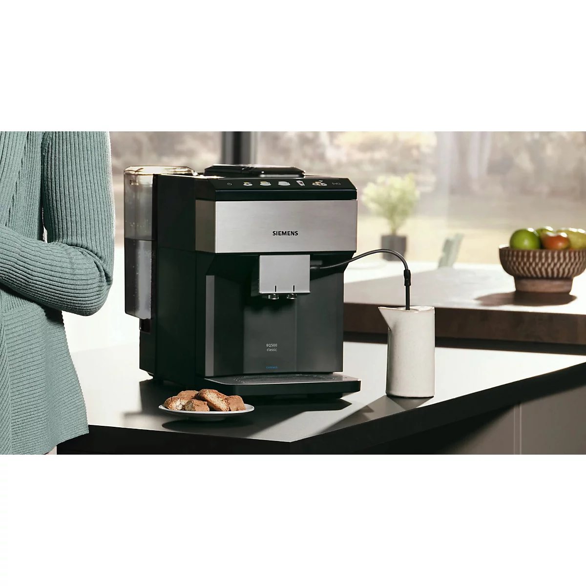 Kaffeemaschine auf Arbeitsplatte, Frau steht daneben, Früchte im Hintergrund.