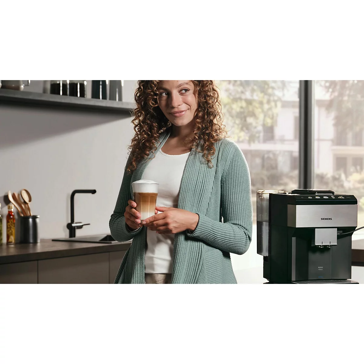 Frau mit lockigem Haar hält Latte Macchiato, lächelnd in der Küche. Kaffeemaschine im Hintergrund.