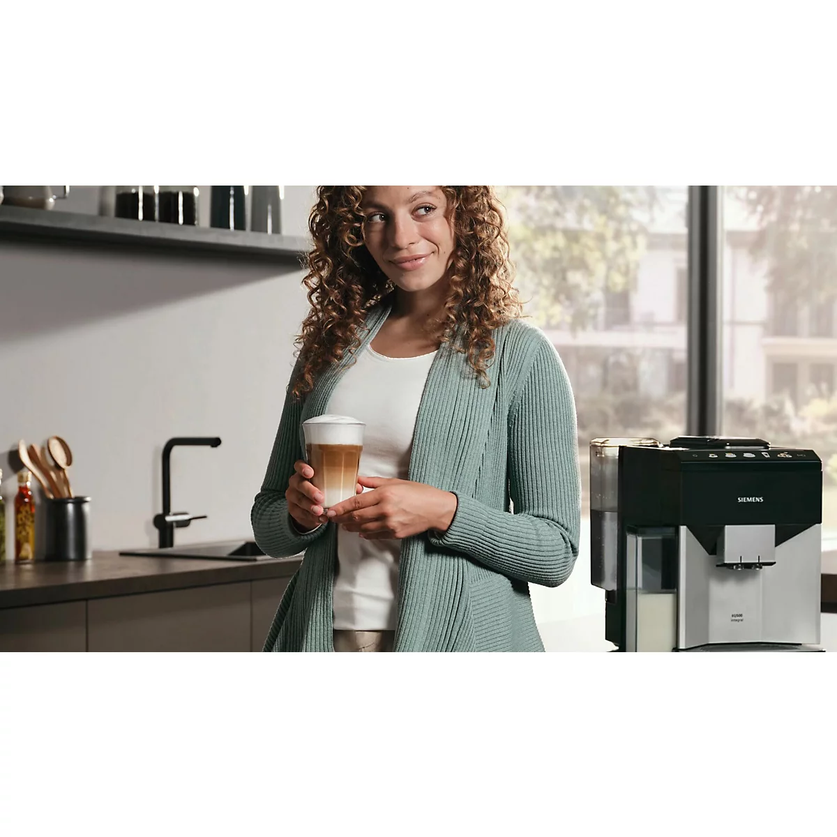 Eine Frau mit lockigem Haar hält einen Latte Macchiato in der Küche. Im Hintergrund eine Kaffeemaschine.