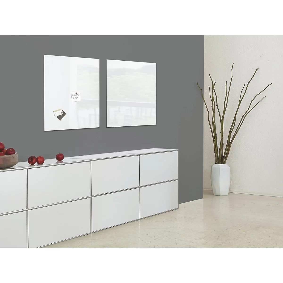 Twee witte glazen borden op grijze muur. Daaronder een wit dressoir, rechts een vaas met takken.