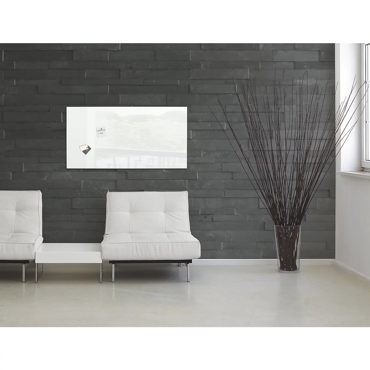 Een wit glazen magneetbord op een donkere stenen muur. Daaronder twee witte fauteuils en een vaas met siergrassen.