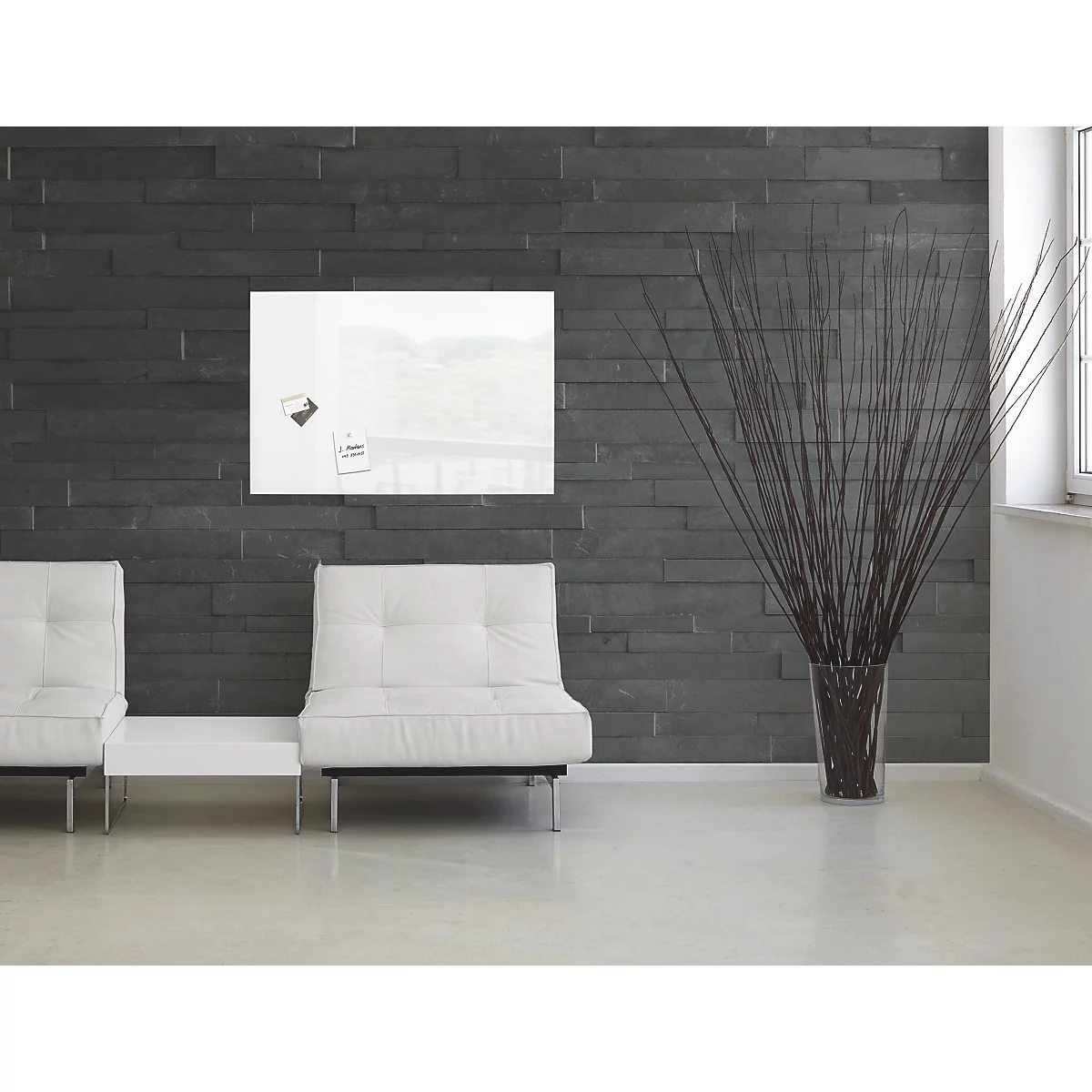 Weisses Glas-Magnetboard an einer dunklen Steinmauer, darunter zwei weisse Sofas. Rechts eine Vase mit Trockenblumen.