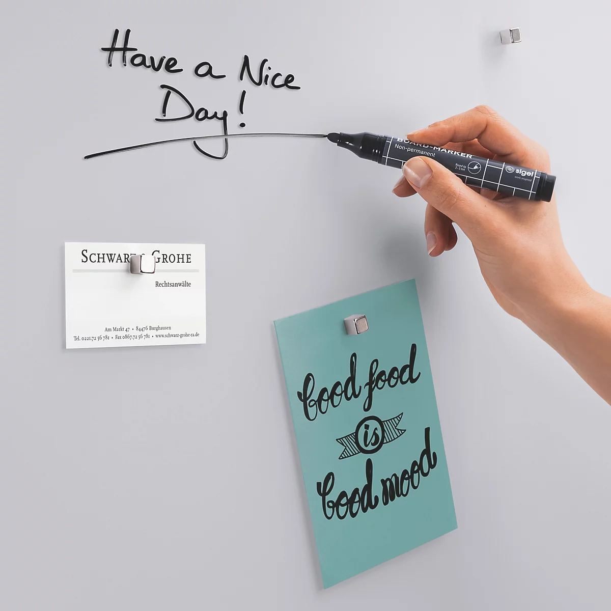 Nahaufnahme einer Hand, die mit einem schwarzen Stift "Have a Nice Day!" schreibt. Auf der Tafel hängen Notizen und eine Visitenkarte.
