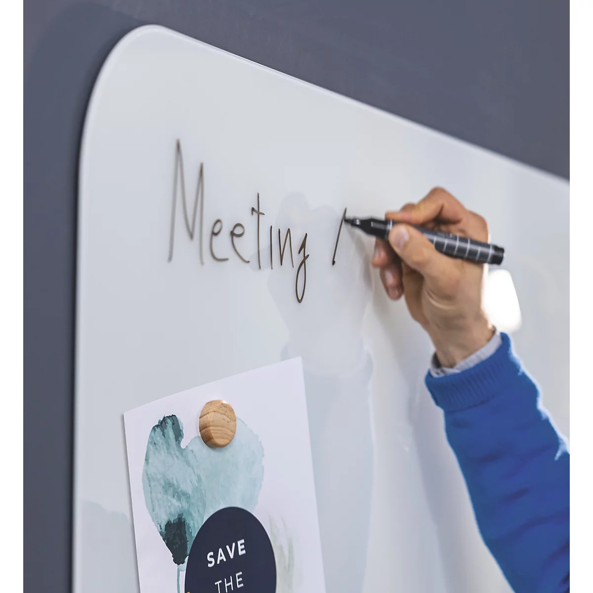 Eine Hand schreibt mit schwarzem Marker das Wort „Meeting“ auf eine weisse Magnettafel. Darunter ein Zettel mit Magnet befestigt.