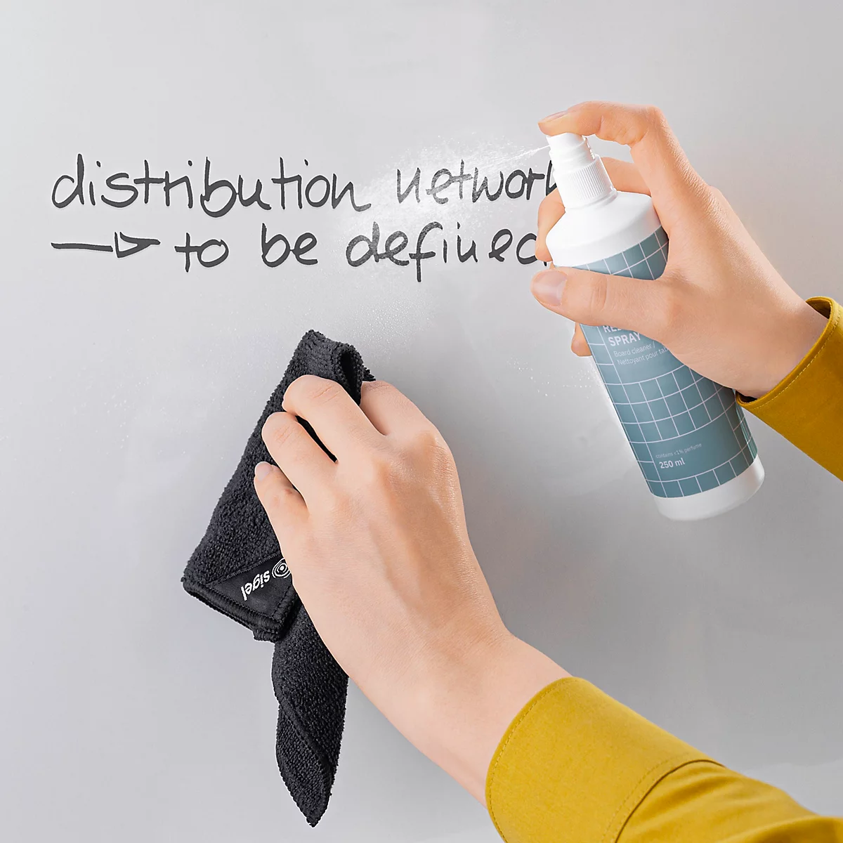 Eine Person besprüht mit einem Reinigungsspray eine Tafel, die mit dem Text "distribution network to be defined" beschrieben ist und wischt diese mit einem schwarzen Tuch ab.