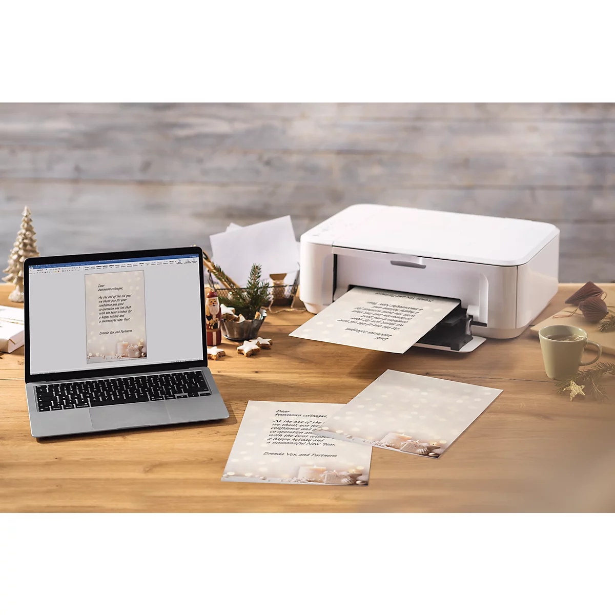 Een laptop en een printer printen kerstkaarten op een houten tafel. Decoraties zoals sterren en een kerstboom zijn op de achtergrond te zien.