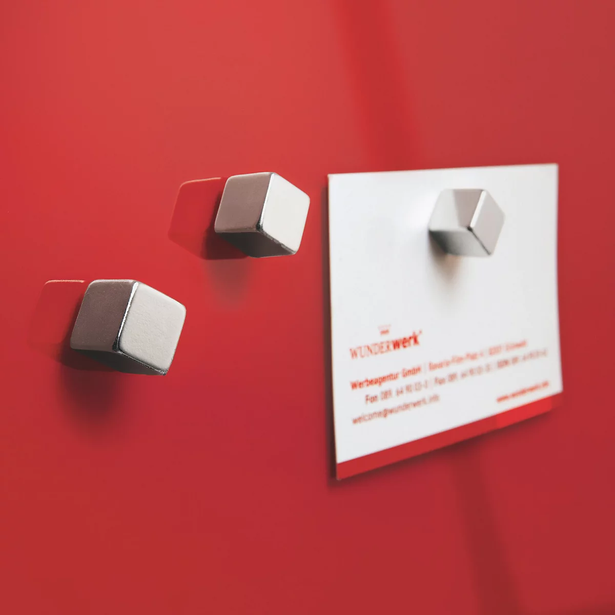 Drei metallische, geometrisch geformte Magnete haften an einer roten Oberfläche, einer hält eine Visitenkarte.