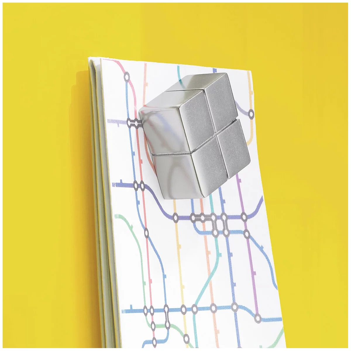 Zilveren kubusvormige magneet op een notitieblok met een metrokaart, tegen een gele achtergrond.