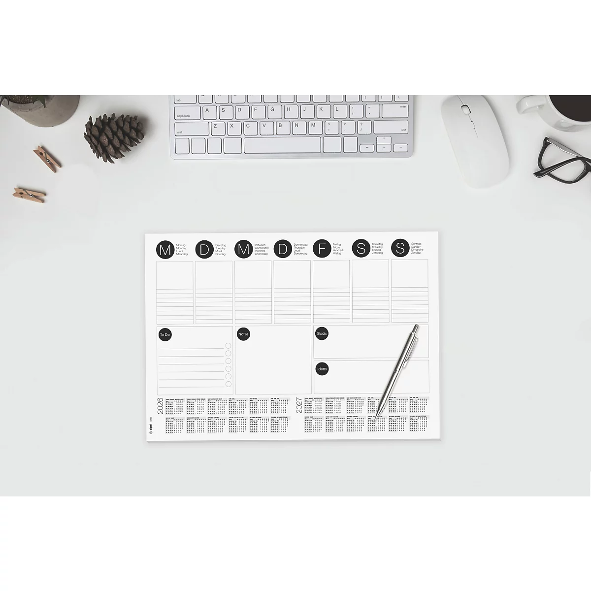 Ein Wochenplaner liegt auf einem weißen Tisch, daneben Tastatur, Maus, Brille, Stift, Tasse und Deko. Der Planer ist weiß mit schwarzer Schrift.
