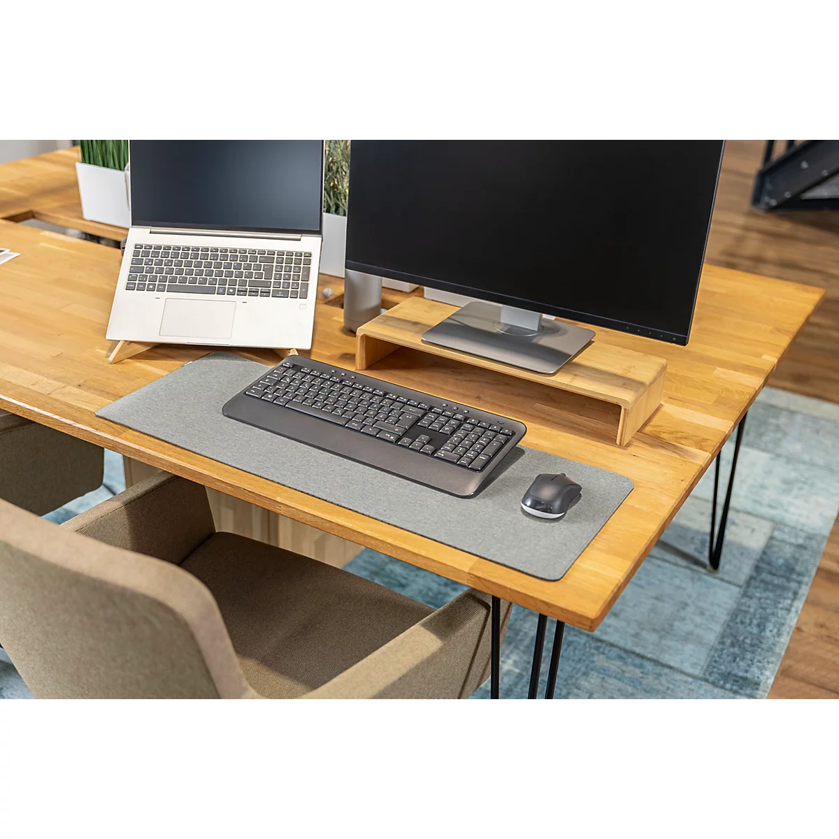 Schreibtisch mit Laptop, Monitor, Tastatur und Maus. Holzplatte, grauer Teppich. Bürostuhl im Vordergrund.