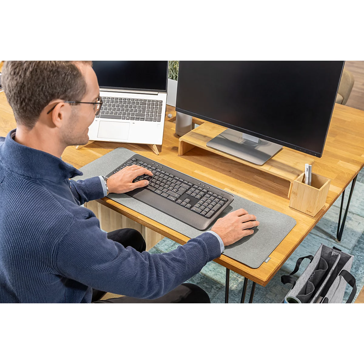 Mann am Schreibtisch mit Brille, tippt auf Tastatur und bedient Maus. Ein Laptop, Monitor und Stifthalter sind ebenfalls zu sehen.