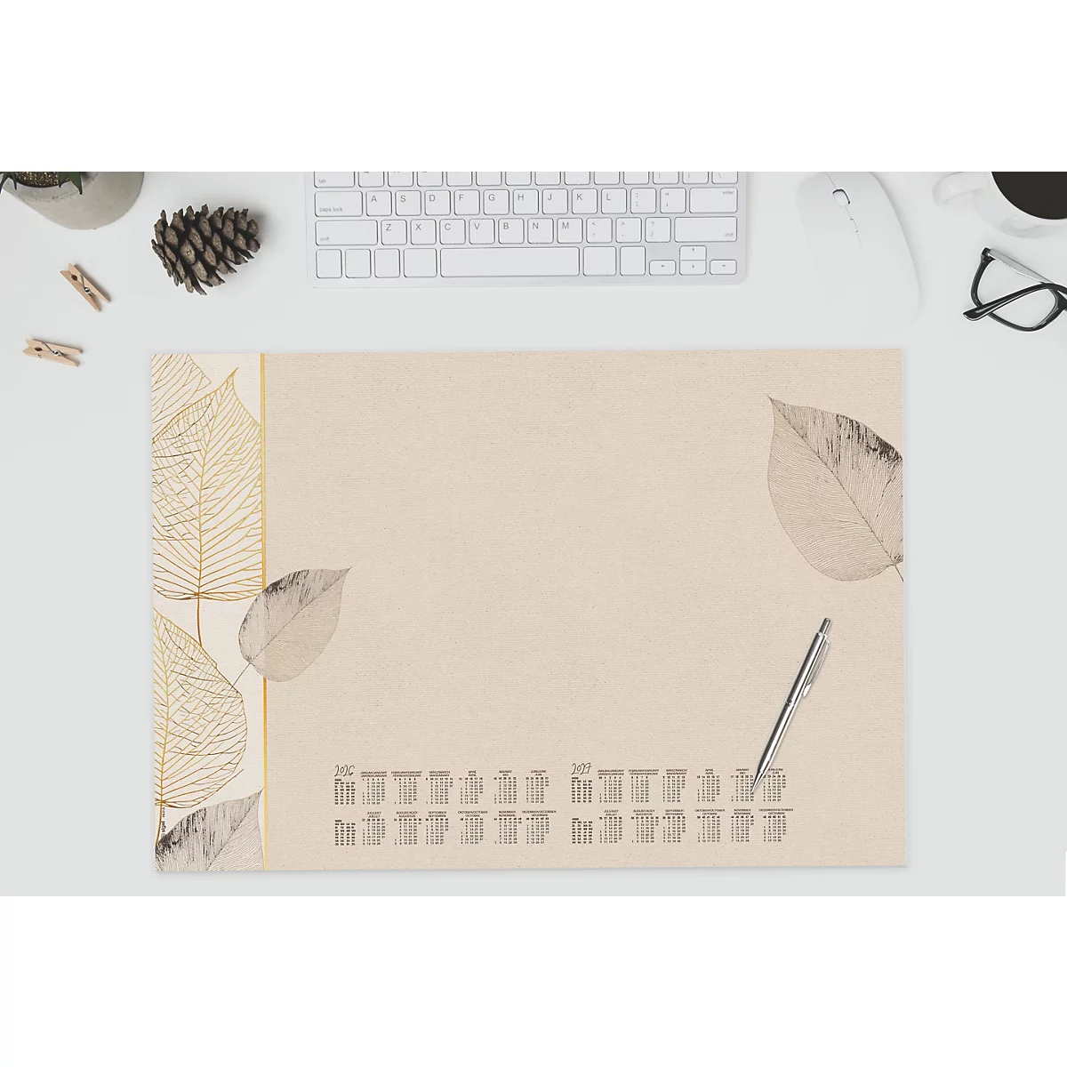 Beige bureau-onderlegger met bladeren en kalender, pen ernaast. Toetsenbord, muis, bril op de achtergrond.