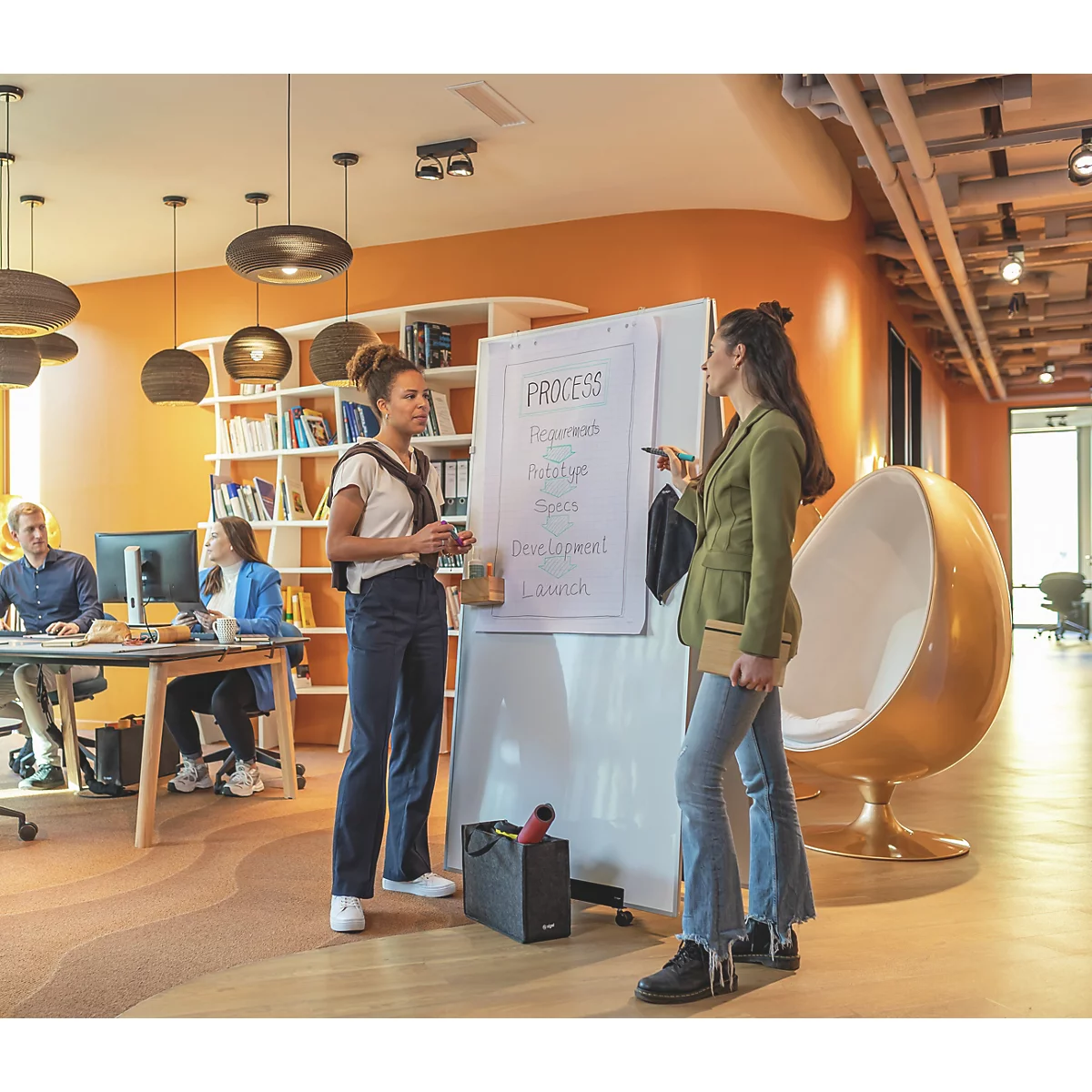 Deux femmes se tiennent devant un tableau blanc avec l'inscription "Process". D'autres personnes travaillent en arrière-plan.