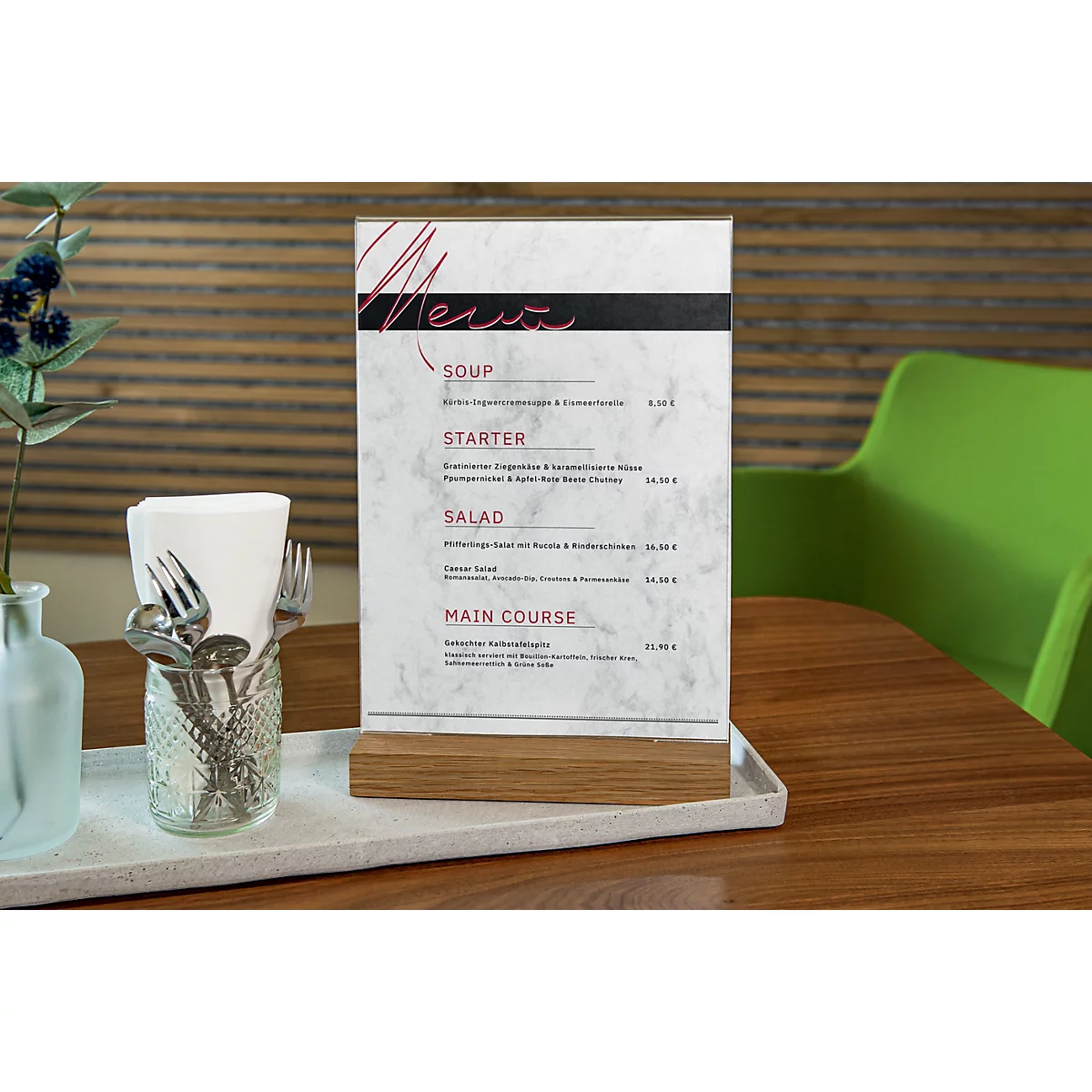 Menu card on a wooden stand. Various cutlery in a glass, white napkin paper and a green chair in the background.