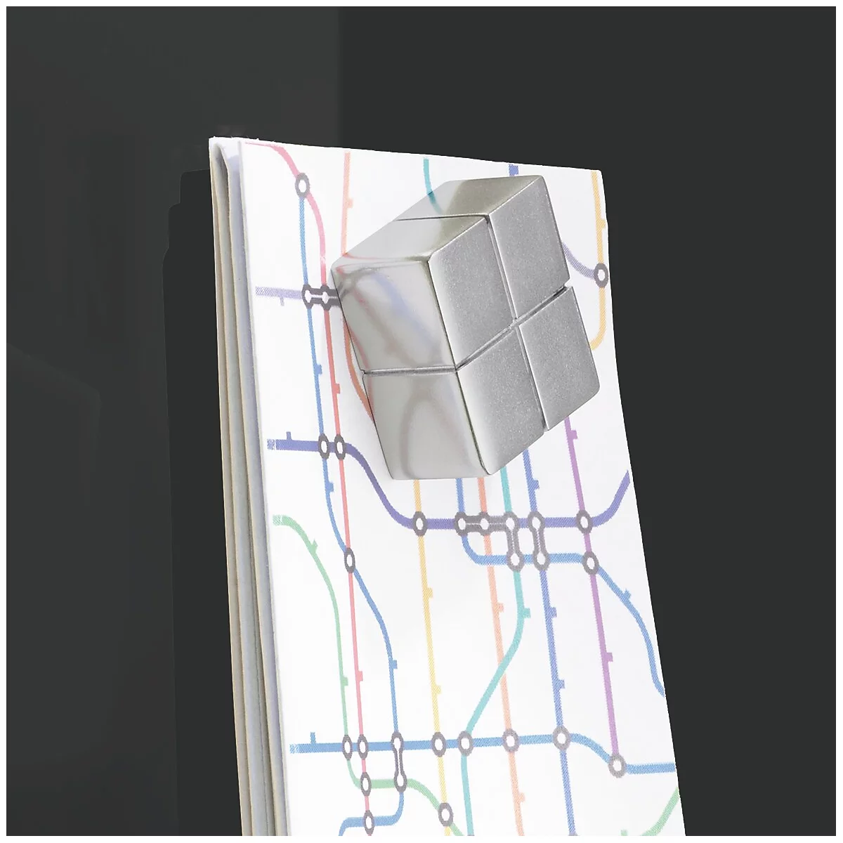 Un aimant cubique argenté est attaché à un bloc-notes avec un motif de lignes de métro. Fond noir.