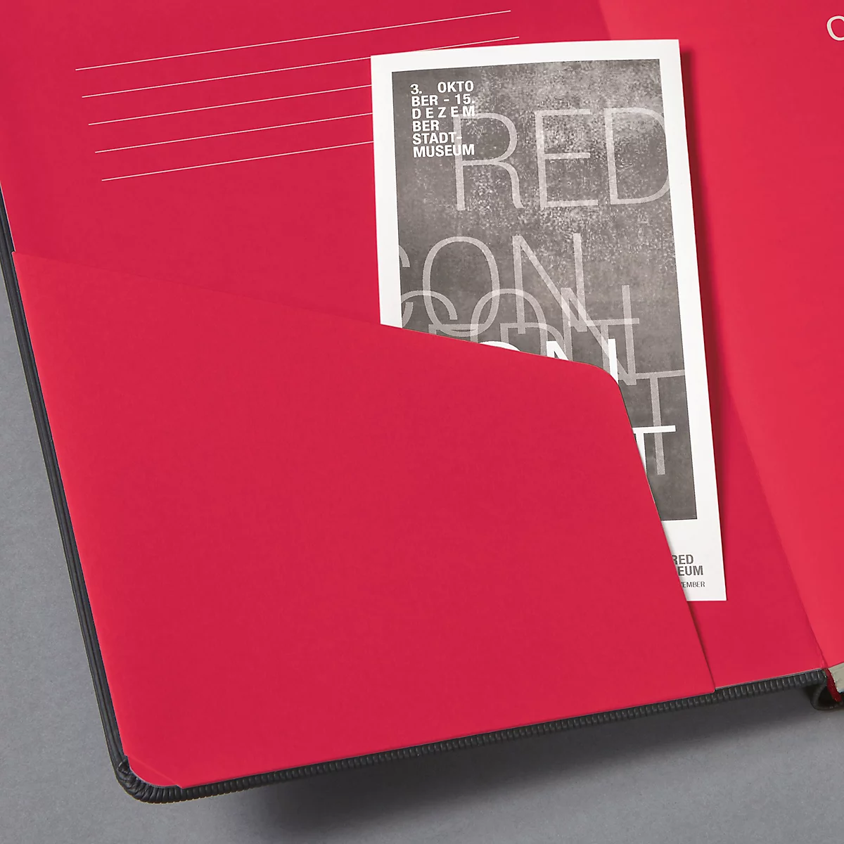 Rotes Notizbuch mit schwarzem Rand. Eine schwarz-weiße Broschüre steckt in einer Tasche.