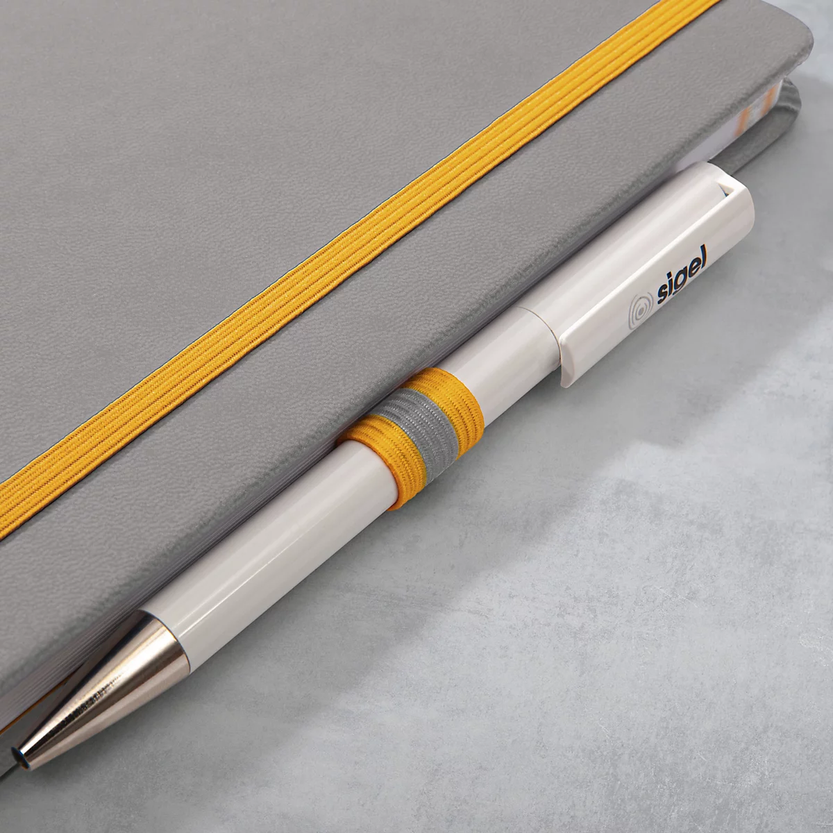 Ein weißer Stift steckt in einem grauen Notizbuch. Der Stift hat einen gelb-grauen Ring und das Logo "sigel".