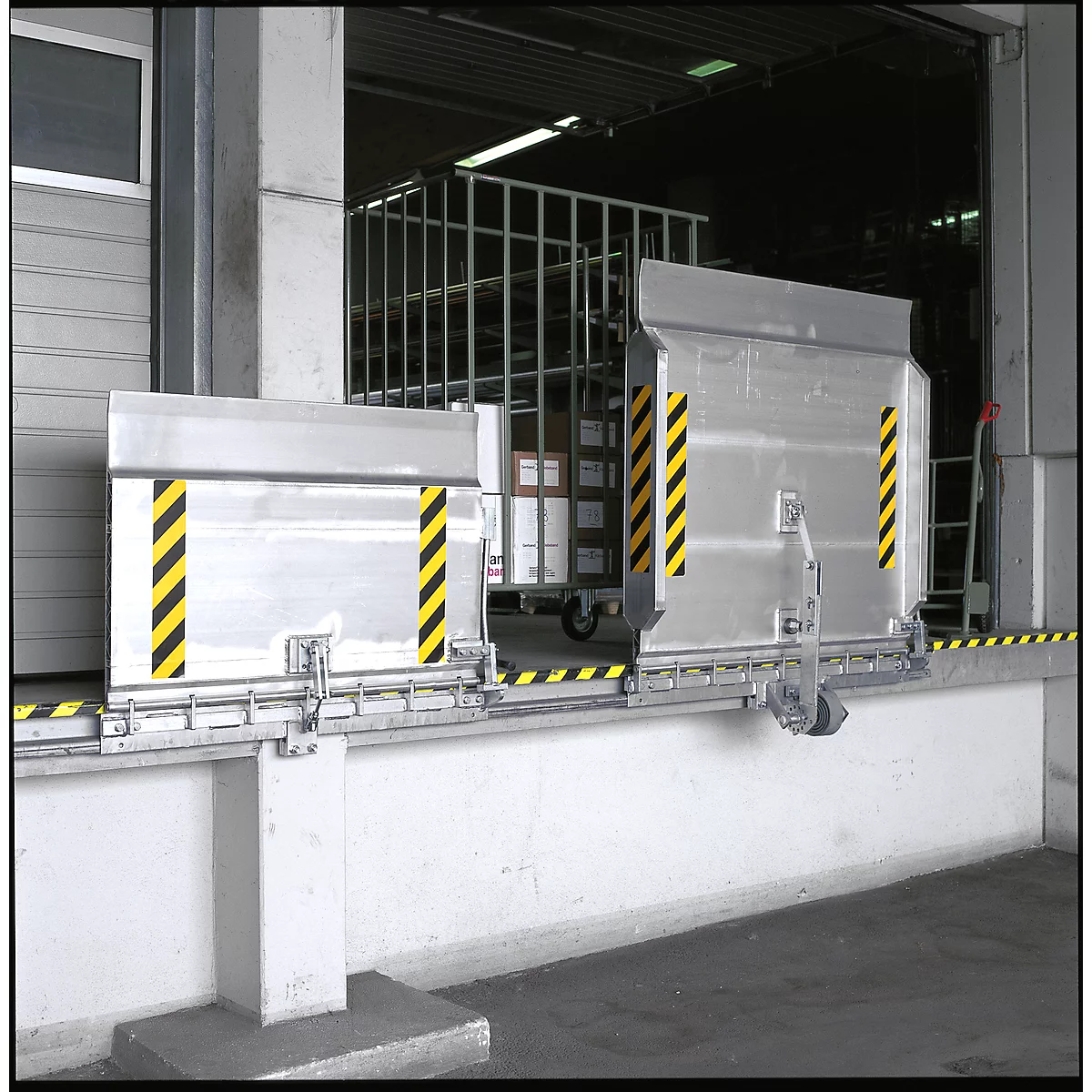 Two silver metal parts, marked yellow and black, in front of a warehouse. In the background, a wire cage cart with boxes.