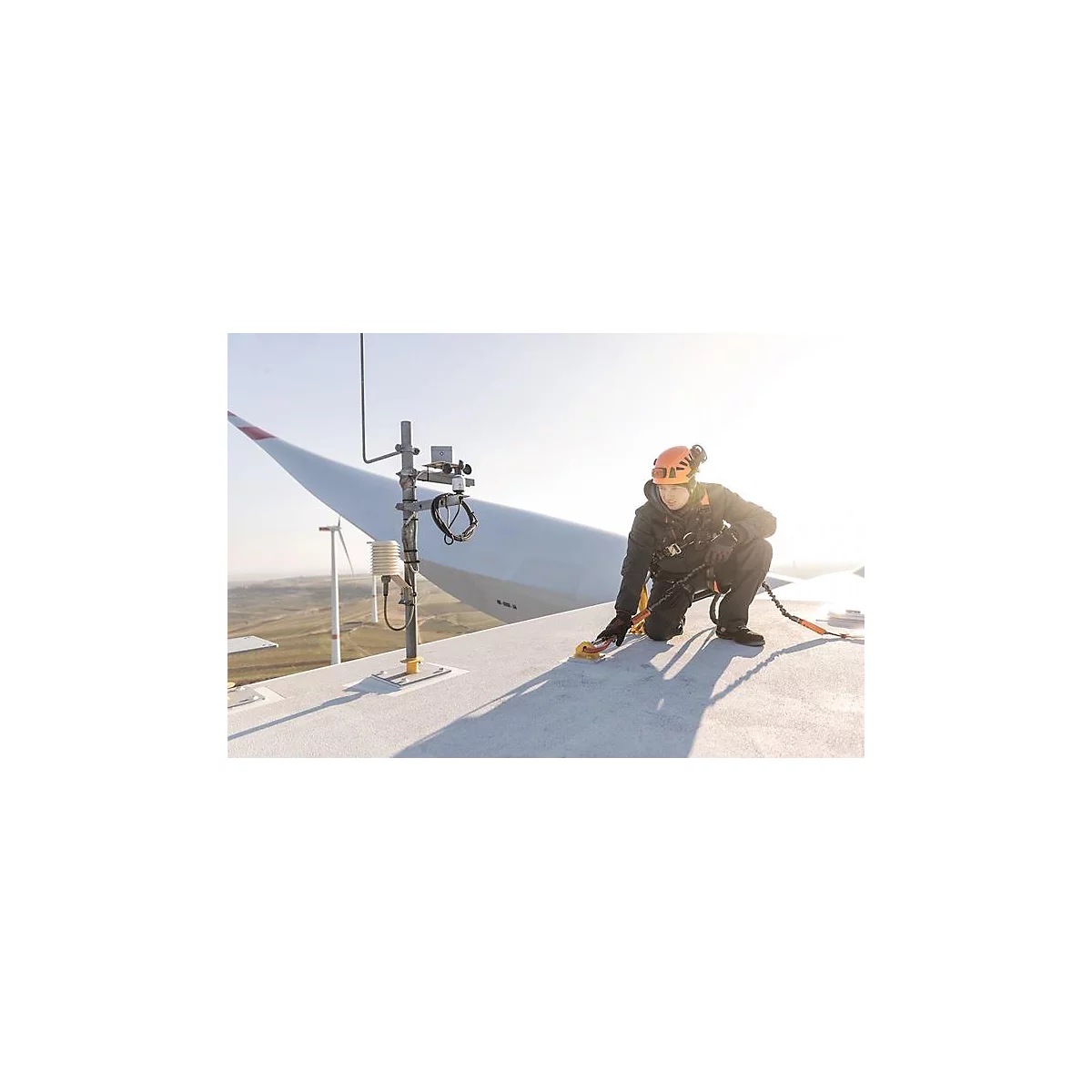 Un travailleur est agenouillé sur une éolienne et travaille. Il porte un casque et est attaché avec une corde.