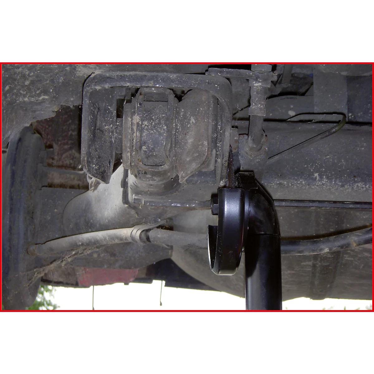 Nahaufnahme der Unterseite eines Autos. Dunkle Metallteile, ein schwarzes Werkzeug und verschiedene Rohre sind zu sehen.