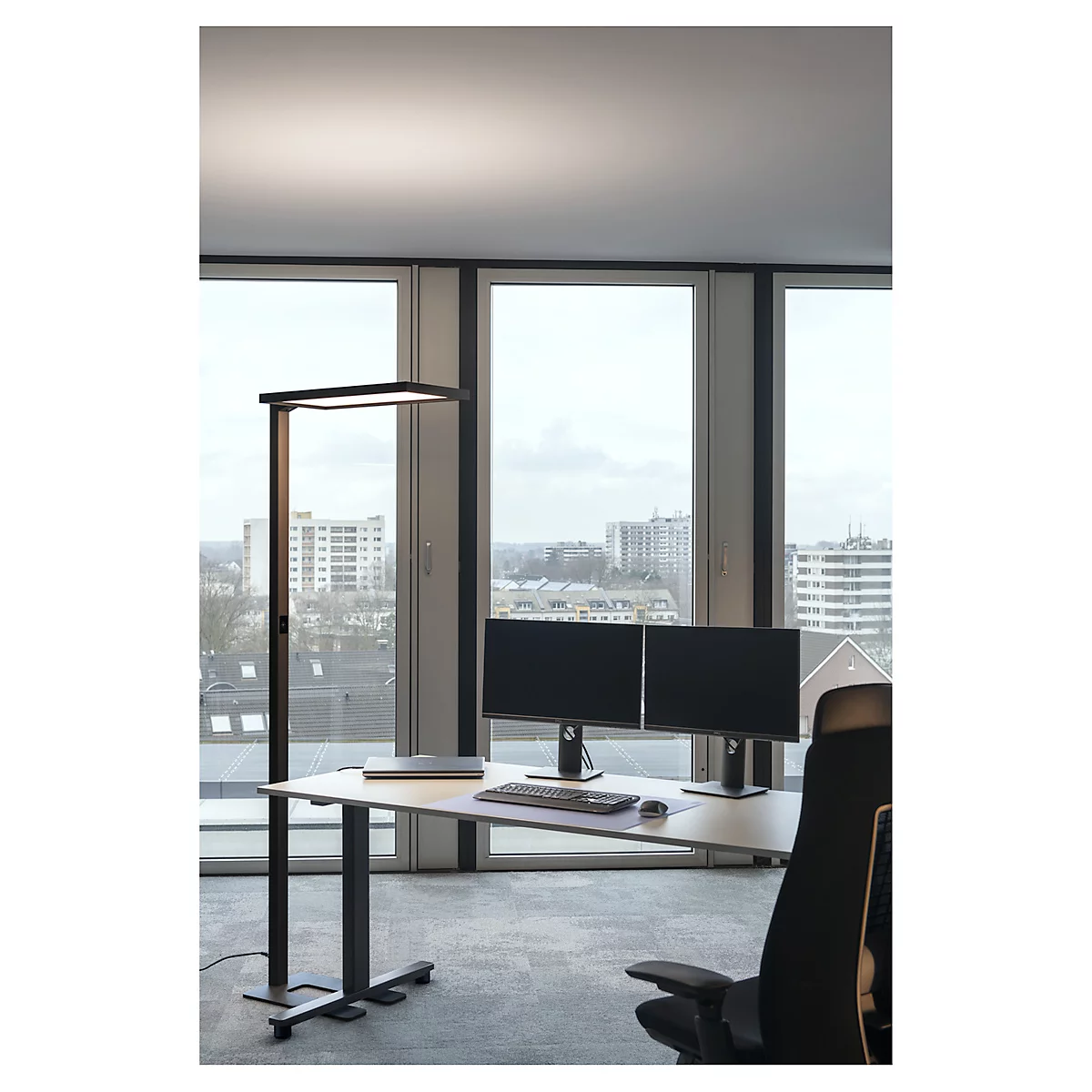 Hohe Stehlampe in einem modernen Büro. Schreibtisch mit zwei Monitoren, Tastatur und Stuhl. Große Fenster mit Stadtblick.