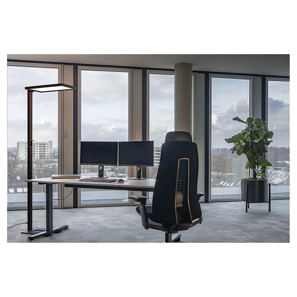 Kantoor met bureau, twee monitoren, bureaustoel, staande lamp en een raampartij met uitzicht.