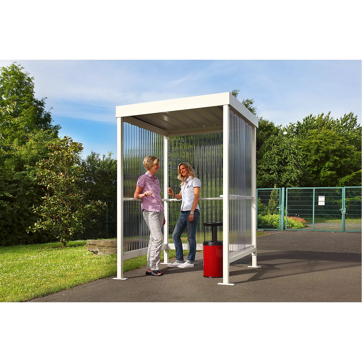 Two women are talking under a white shelter with transparent side walls. A red trash can stands next to it.