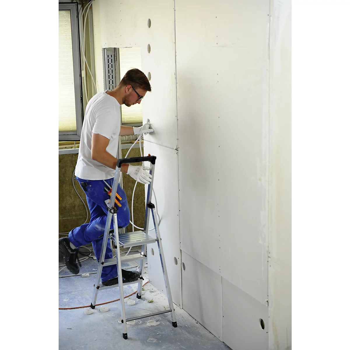 A man on a ladder attaching wires to a wall in a shell building. He wears glasses, a white T-shirt and blue work pants.