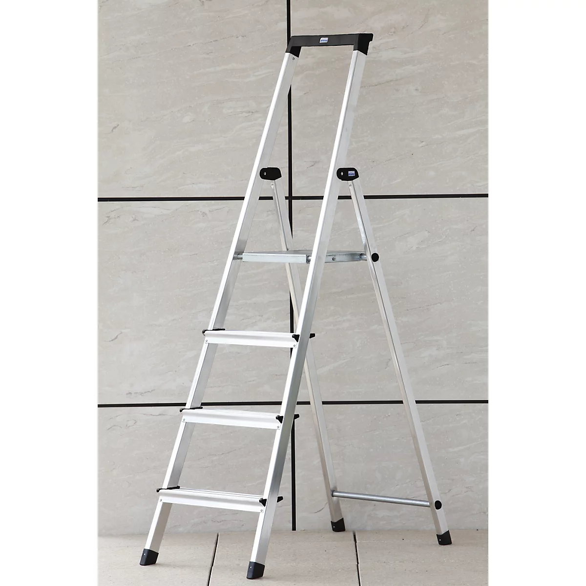 A silver step ladder stands in front of a light gray wall. Four steps, black feet.