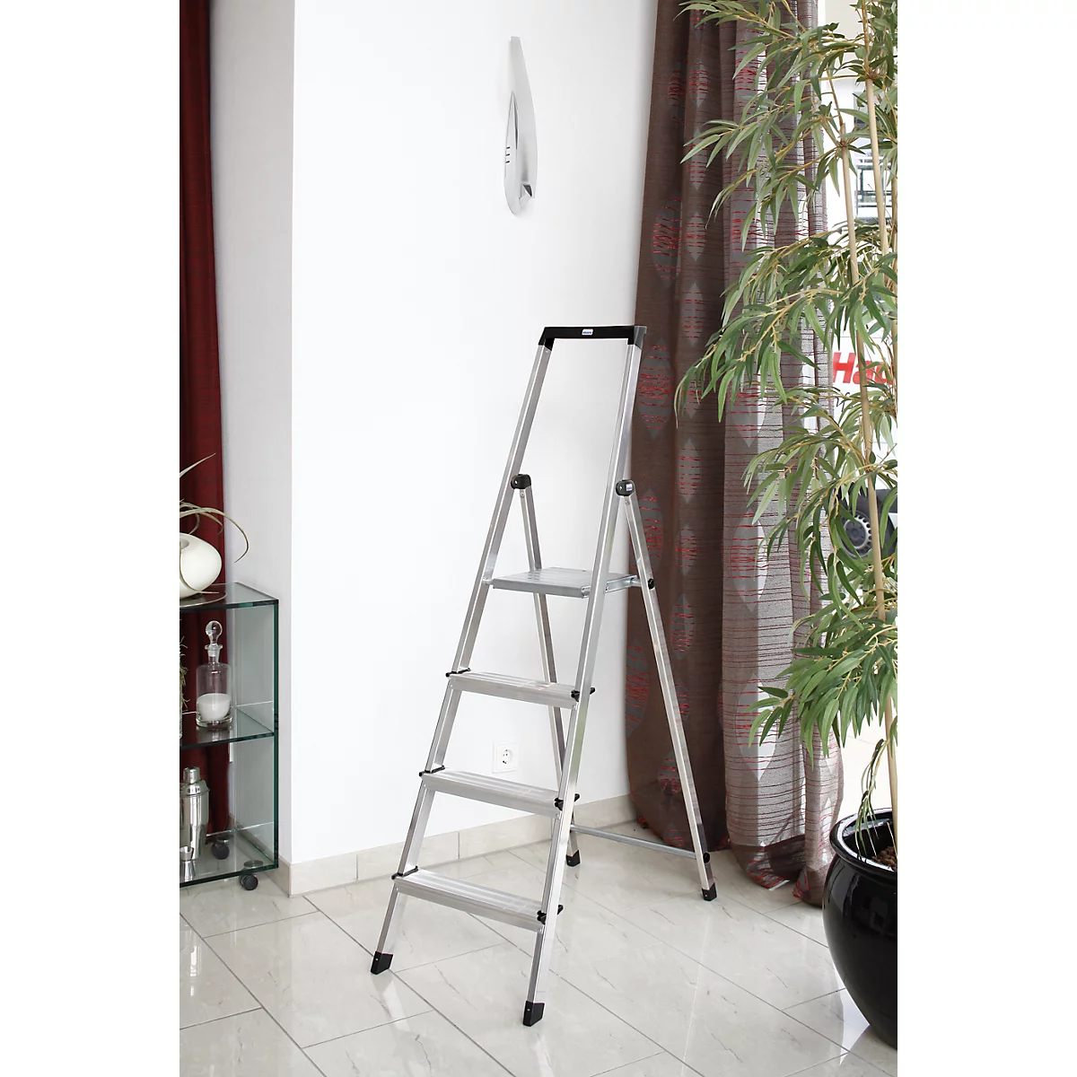 Aluminum step ladder against a white wall. A shelf on the left, a plant on the right. A decorative object hangs on the wall.