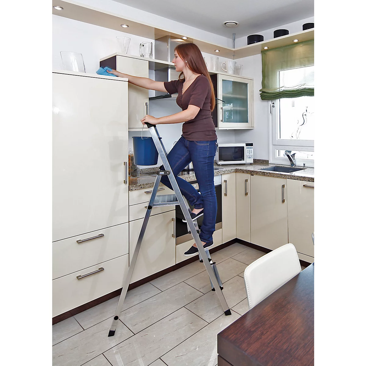 Woman cleaning a top cabinet in the kitchen using a step ladder.