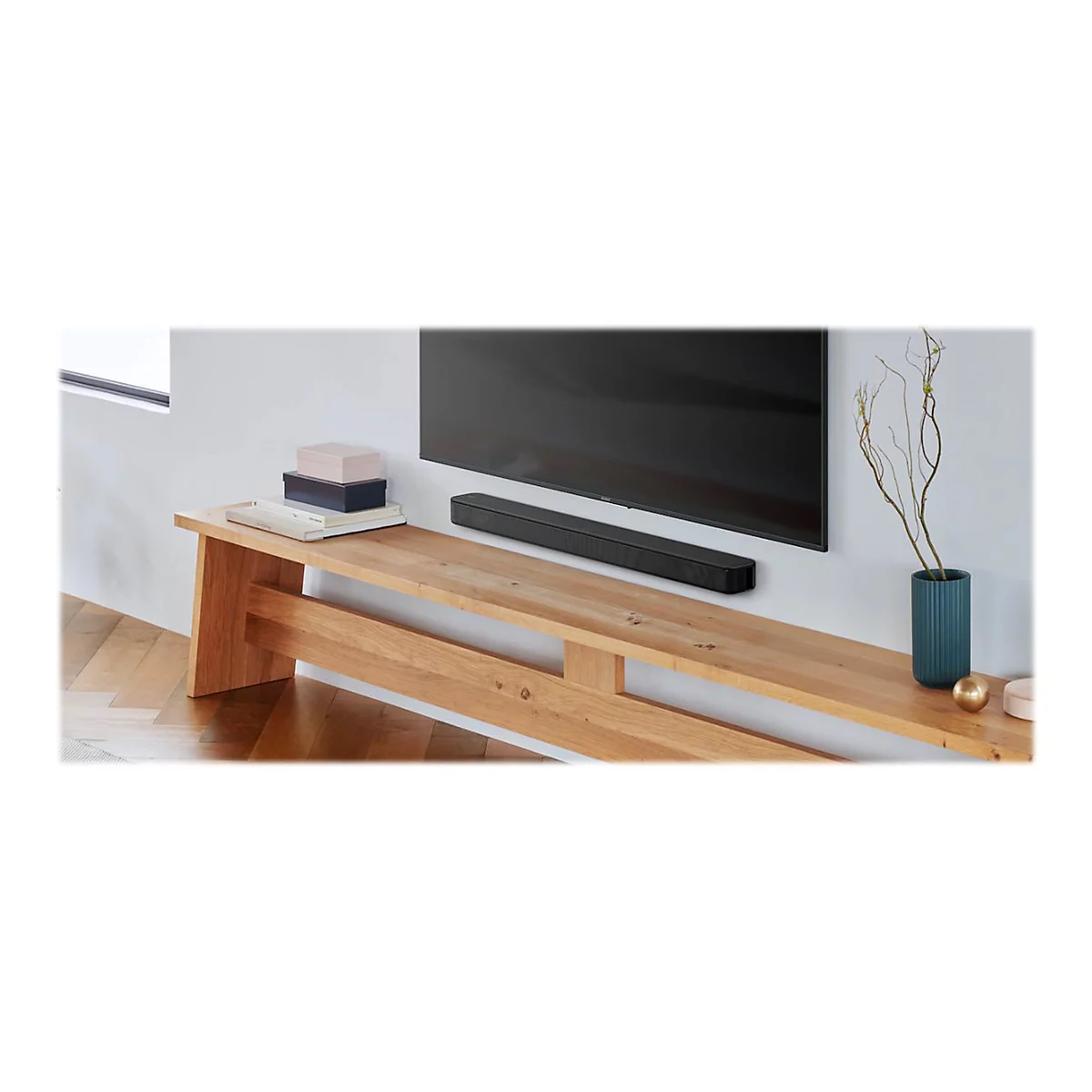 Eine schwarze Soundbar unter einem Fernseher an einer Holzwand. Auf einem Holzregal stehen Deko-Gegenstände.