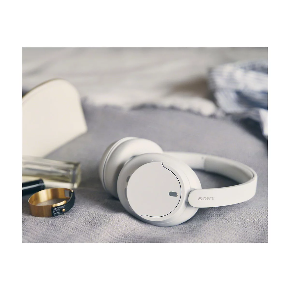 Weiße Kopfhörer von Sony, liegen neben Schmuck und einem Kosmetikartikel auf einer grauen Decke.