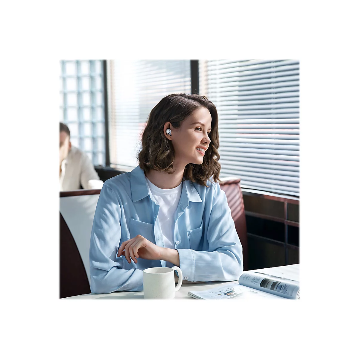 Eine lächelnde Frau mit Kopfhörern sitzt in einem Café. Sie trägt ein blaues Hemd, die Sonnenblenden sind im Hintergrund zu sehen.