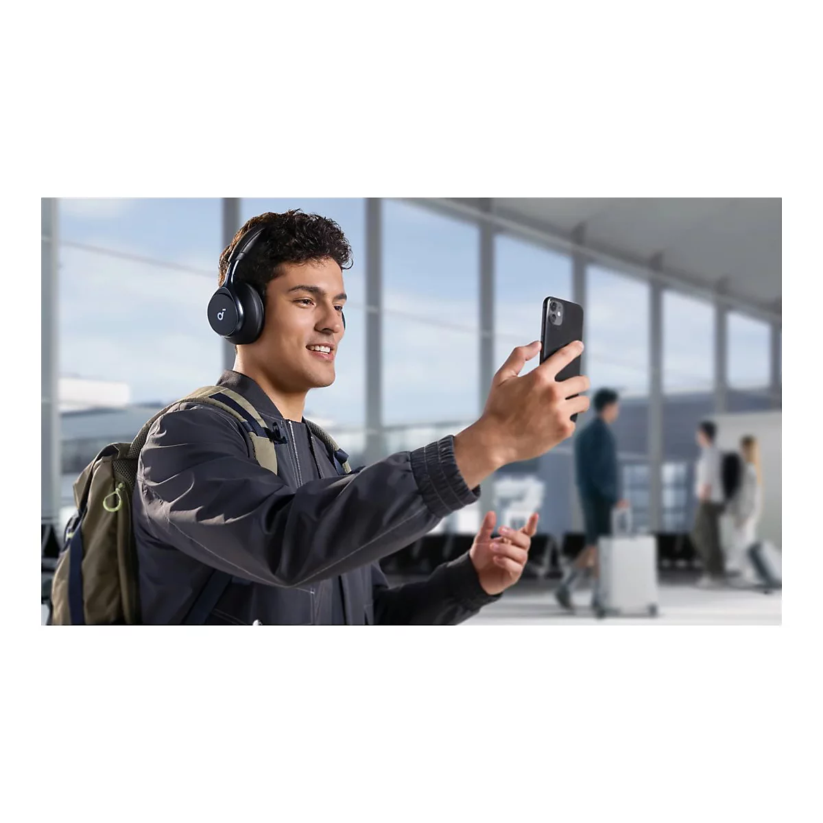 Ein Mann mit KopfhÃ¶rern macht ein Videoanruf auf seinem Smartphone, lÃ¤chelnd in einem Flughafen. Er trÃ¤gt einen Rucksack.