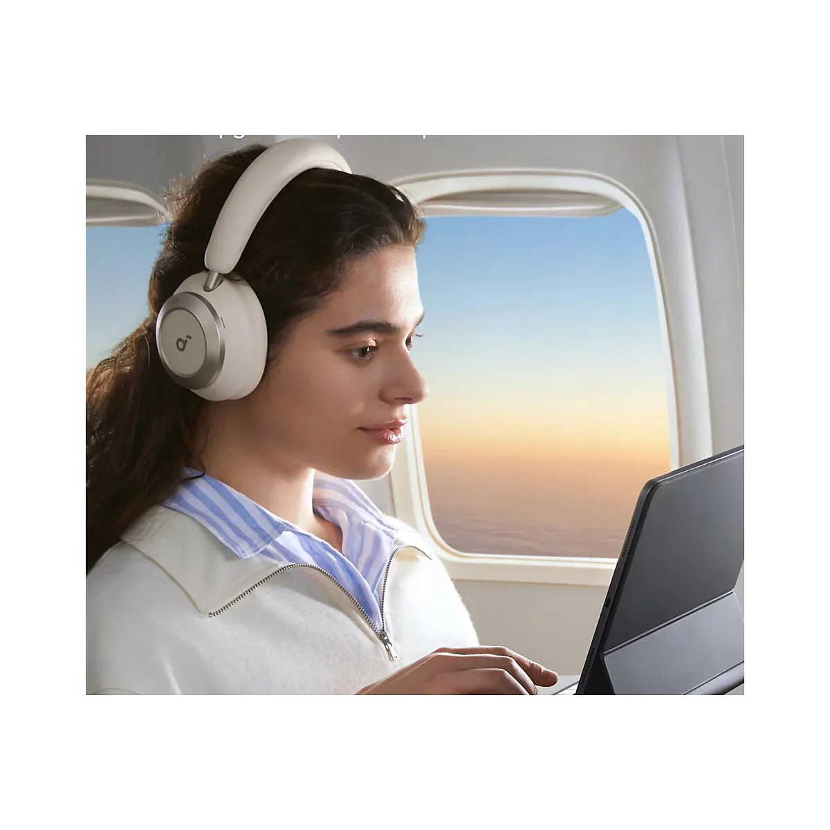 Junge Frau im Flugzeug, mit weiÃŸen KopfhÃ¶rern und Laptop am Fenster. Blick auf den Sonnenaufgang.