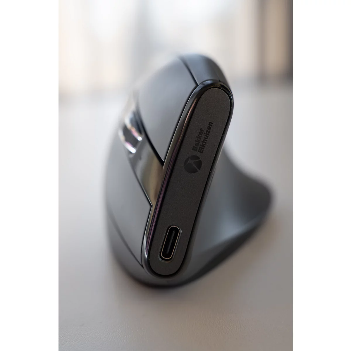 Souris ergonomique grise avec texte "Bakker Elkhuizen". Port USB-C, posé sur une surface claire.