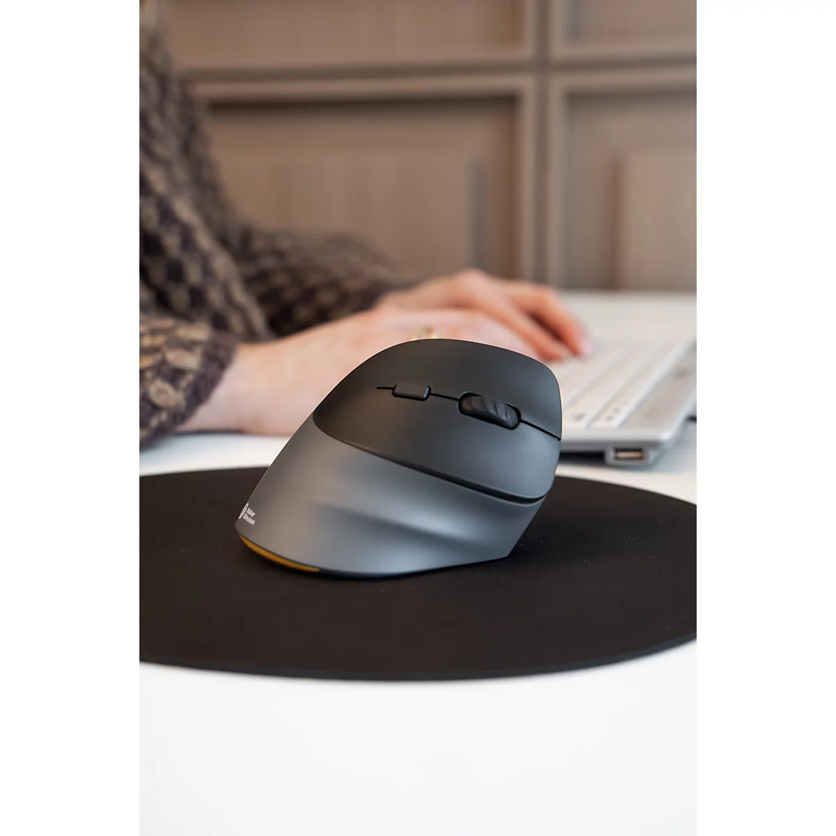 Une souris verticale ergonomique gris foncé posée sur un tapis de souris noir devant une personne qui travaille.